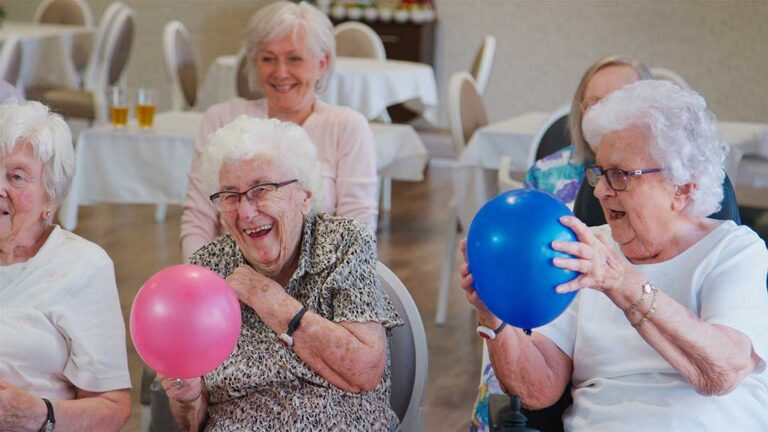 Older adult arranging family photographs and a favorite chair in a bright assisted living apartment.